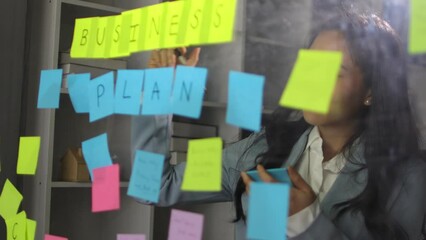 Creative young Asian woman thinking and planning a startup business with paper stickers on a transparent board.