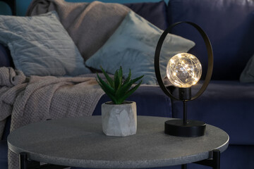 Glowing lamp and houseplant on coffee table near blue sofa in dark living room