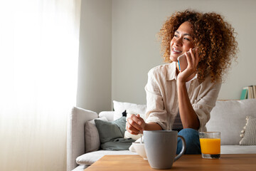 Young multiracial hispanic woman talking on the phone while having breakfast sitting on the sofa....