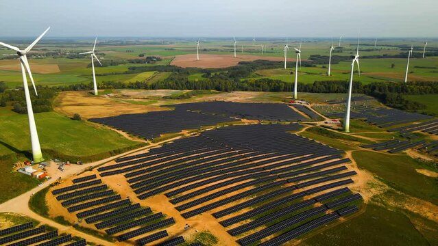 Aerial footage of solar panels plant and wind turbines in a wind farm generating green electric energy on a wide green field on a sunny day, in Taurage, Lithuania, zooming out