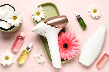 Modern photoepilator with bottles of cosmetic products and beautiful flower on pink tile background