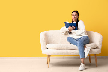 Fototapeta na wymiar Beautiful young woman with book sitting on comfortable sofa near yellow wall