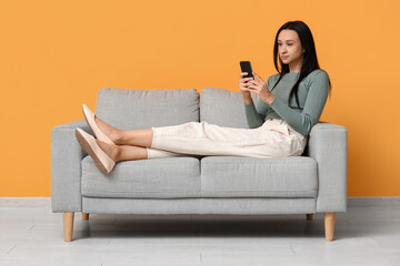 Obraz premium Beautiful young woman with mobile phone sitting on comfortable sofa near yellow wall