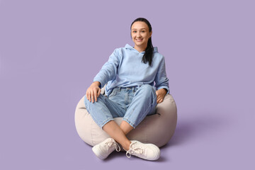 Beautiful young woman relaxing on comfortable beanbag chair against purple background