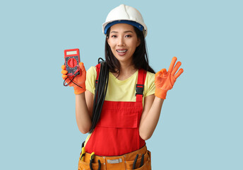 Young female electrician with multimeter showing OK gesture on blue background