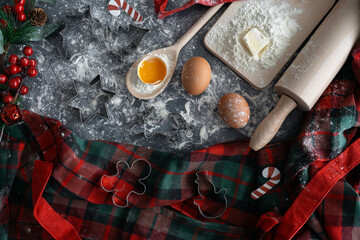 Christmas cookies baking composition on a flour-dusted kitchen table. Wooden utensils, fresh dough ingredients, tartan plaid and cookie cutters for cutting out shapes. Holiday season winter flat lay.