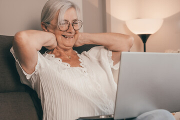Relaxed mature senior woman sitting on sofa using laptop technology, enjoying carefree moments...