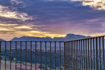 view of a sunset in a mountain landscape with dramatic and colorful cloudy skies seen from a lookout point with a metal railing