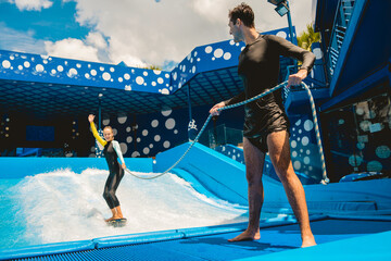 Beautiful young woman surfing with trainer on a wave simulator at a water amusement park