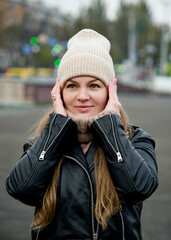 beautiful smiling girl with long blonde hair staying in the street in beige handmade knitted hat and black leather jacket