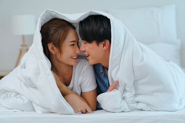 Young Asian couple lovers use noses touch together with love, smiling faces and happiness in bed while using blanket cover heads.