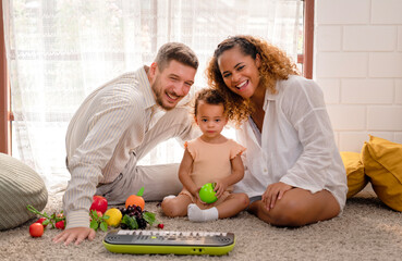 Portrait of happy family plays with kid in living room with diversity fruit toys on the floor at home, relaxing time to play with little child for developing remember brain, muscles, colors and focus
