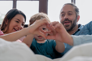 Parent and little kid making hands heart together while playing on bed in bedroom at home with smiling face and happy moment, happiness family. Father, mother, boy play together to develop muscles
