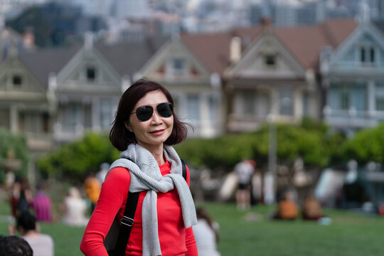 Senior woman traveler with red coat and sunglasses traveling alone to visit painted lady houses in San Francisco, shooting image while standing on hills with houses background, vacation, trip, holiday