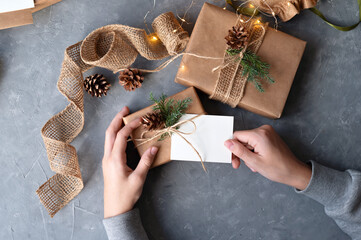 Preparing Christmas holiday presents, brown gift boxes decorated with pine cones and twigs, greeting postcard or tag and present box in hands. Winter holiday celebration, card mockup with copy space