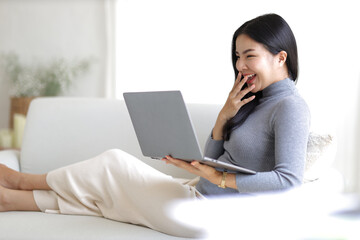 Young asian woman using computer laptop while seated on couch at home. Happy girl relaxing comfortable, using laptop in the living room, spending lazy weekend, watching movie, shopping online.