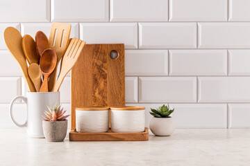 Ceramic white jars, wooden spoons in potstav and wooden cutting board on kitchen light countertop. Front view. A copy of the space. Eco style.