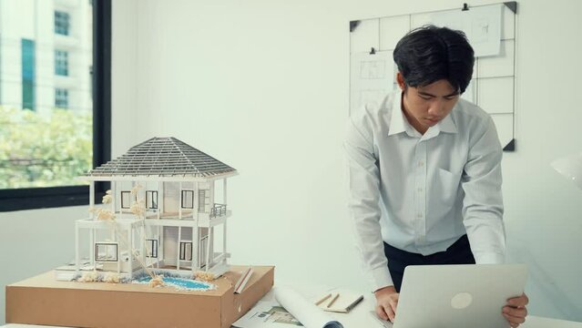 Close up of professional male architect engineer measure house model and using laptop analysis data at meeting table with house model and map . scatter around. Focus on hand. Immaculate.