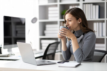 Attractive woman drinking coffee while working on laptop in office at home in morning.