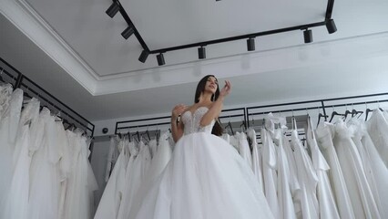 A happy young woman at the fitting of a wedding dress in a wedding salon, she rotates from side to side standing against the background of many wedding dresses on hangers.
