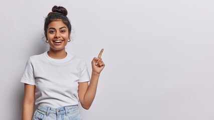 Horizontal shot of pretty Iranian woman with hair bun dressed in casual t shirt and jeans points index finger on copy space for your advertisement isolated on white background. Look at this promotion