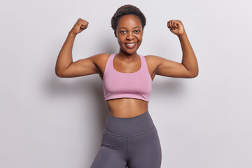 Look how I can do. Dark skinned African sportswoman raises arms and shows biceps leads healthy lifestyle dressed in activewear isolated over white background. Sport is life. Female strength.