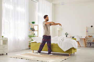 Man in pyjamas sleepwalks at home. Man is like sleepy zombie not fully awake from dream in early morning. Man with sleep disorders is walking with closed eyes in living room in his house or apartment - obrazy, fototapety, plakaty