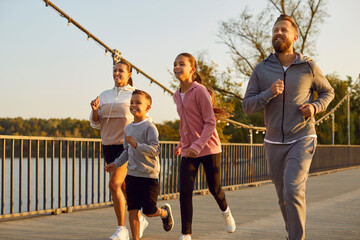 Happy active family in sportswear running together in park along a bridge having sport workout...