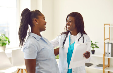 Portrait of smiling friendly female african american doctor therapist talking with a young woman patient holding report file with appointment standing in office. Medicine and health care concept. - obrazy, fototapety, plakaty