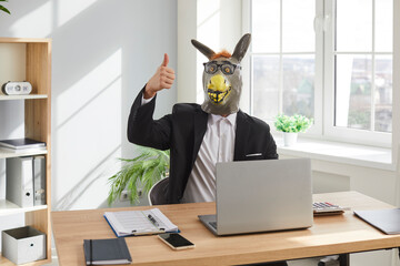 Funny business man and company employee wearing suit and animal donkey mask sitting at the desk...