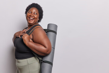Studio shot of cheerful obese African woman with curly hair laughs positively keeps hand on chest carries rubber fitness mat dressed in active wear goes in for sport to loose weight isolated - 680467569