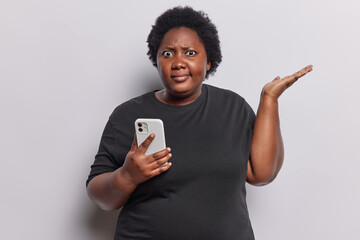 Confused overweight curly woman shrugs shoulders holds smartphone has connection error annoyed with discharged or broken cellphone dressed in casual black t shirt isolated over white background - obrazy, fototapety, plakaty