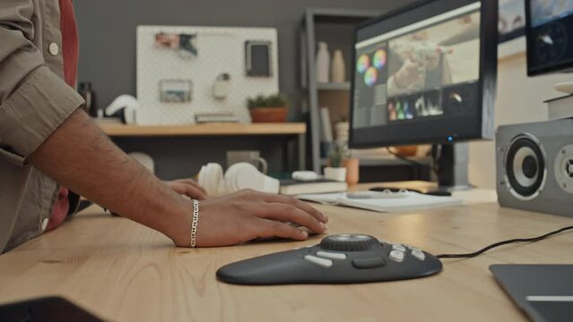 Close-up shot of hands of anonymous male designer standing at desk in office or at home and working on video clip in editing software on computer, using professional multimedia controller