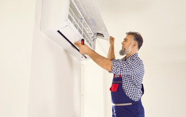 Fototapeta na wymiar Professional electrician worker man with screwdriver maintaining, cleaning modern air conditioner indoors. Mature gray-haired technician in white room repairing or installing air conditioner.