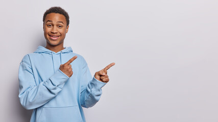 Dark skinned African man points both index fingers aside shows place for your advertisement wears casual comfortable blue sweatshirt isolated over white background. Hey you should check this out - obrazy, fototapety, plakaty