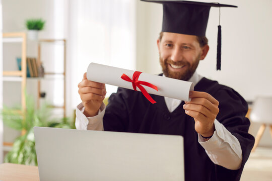 Happy smiling adult male student in graduation hat and gown sitting at desk with laptop notebook PC at home and holding paper diploma scroll in hands. Online degree, remote higher education concept