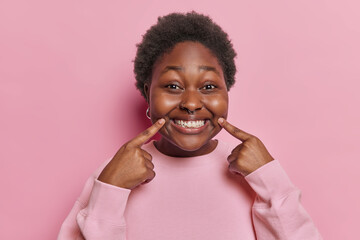 Photo of positive dark skinned plump lovely woman points index fingers at toothy smile shoows white perfect teeth after visiting dentist dressed in casual jumper isolated over pink background