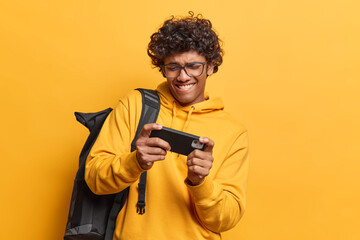 Addicted curly haired teenage Hindu man playing mobile video games online on cellphone bites lips dressed in casual sweatshirt carries rucksack isolated over yellow background uses his gadget