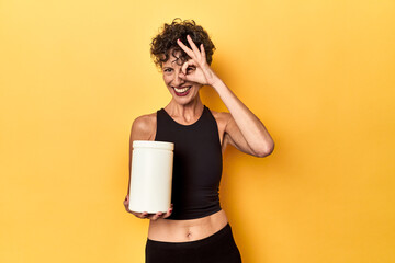 MIddle aged athlete woman holding protein supplement on yellow excited keeping ok gesture on eye.