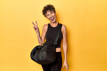Sportswoman with gym backpack on yellow studio joyful and carefree showing a peace symbol with...