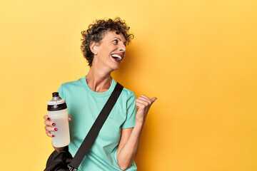 Sportswoman with gym bag & water on yellow studio points with thumb finger away, laughing and...