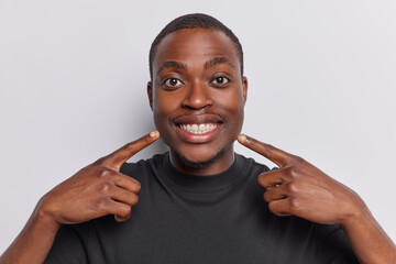 Young positive dark skinned man points at toothy smile shows perfect teeth dressed in casual black t shirt attracts your attention to mouth dressed in casual black t shirt isolated on white background - obrazy, fototapety, plakaty