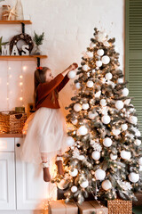 Merry Christmas and Happy Holidays. Cute little child girl is decorating the Christmas tree indoors. The morning before Xmas.