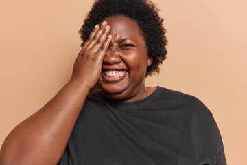 Indoor shot of overweight dark skinned woman covers half of face with palm smiles broadly dressed...