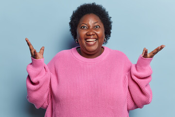 Indoor shot of chubby dark skinned woman keeps palms raised up exclaims loudly shows white teeth...