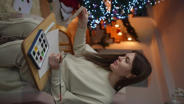 Vertical video. A woman, having painted a Christmas wreath on a postcard in watercolor, positively evaluates her creativity while sitting in a rocking chair against the backdrop of a Christmas tree