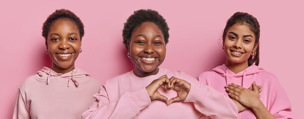 Set of cheerful dark skinned woman smiling gladfully happy chubby African female making heart gesture and expressiing love dark haired Iranian girl demonstrates gratitude gesture isolated on pink wall