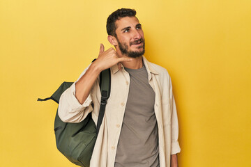 Young Hispanic man with travel backpack showing a mobile phone call gesture with fingers.