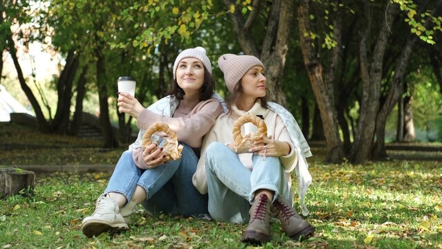 Two cute women in warm clothes sit hugging in an autumn park. Girlfriends enjoying beautiful weather, drinking tea or coffee, eating fresh bagels from the bakery - Powered by Adobe