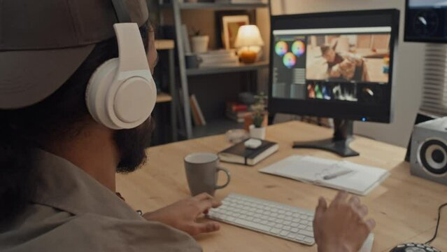 Rear close-up shot of young Middle Eastern man with ponytail and beard working on photos or footage in editing software at home, and putting on wireless headphones to listen to music